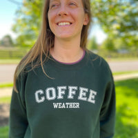 Load image into Gallery viewer, Coffee Weather crewneck sweatshirt in forest green on woman in a park, white varsity-style design, cozy coffee crewneck