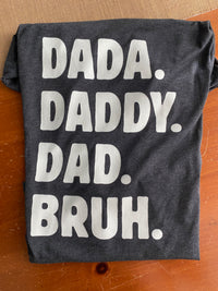 Load image into Gallery viewer, Dada. Daddy. Dad. Bruh. Everyday Tee in dark grey heather, flat lay on wood surface