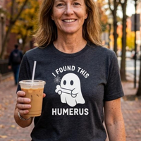 Load image into Gallery viewer, A woman outdoors wearing a dark charcoal heather graphic T-shirt with a white ghost and the text 'I FOUND THIS HUMERUS' on the front, while holding an iced coffee.